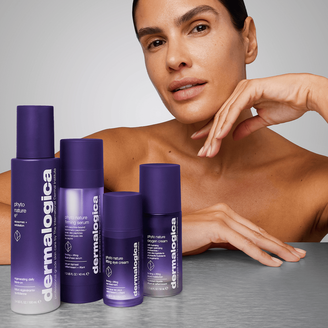Eine Person posiert hinter vier Dermalogica Phyto Nature E2 Exosom Treatment-Produkten mit violetter Verpackung und weißem Text, die auf einer grauen Fläche ausgestellt sind - und die fortschrittlichen Leave-on-Formeln hervorheben, die die Hautregeneration unterstützen.