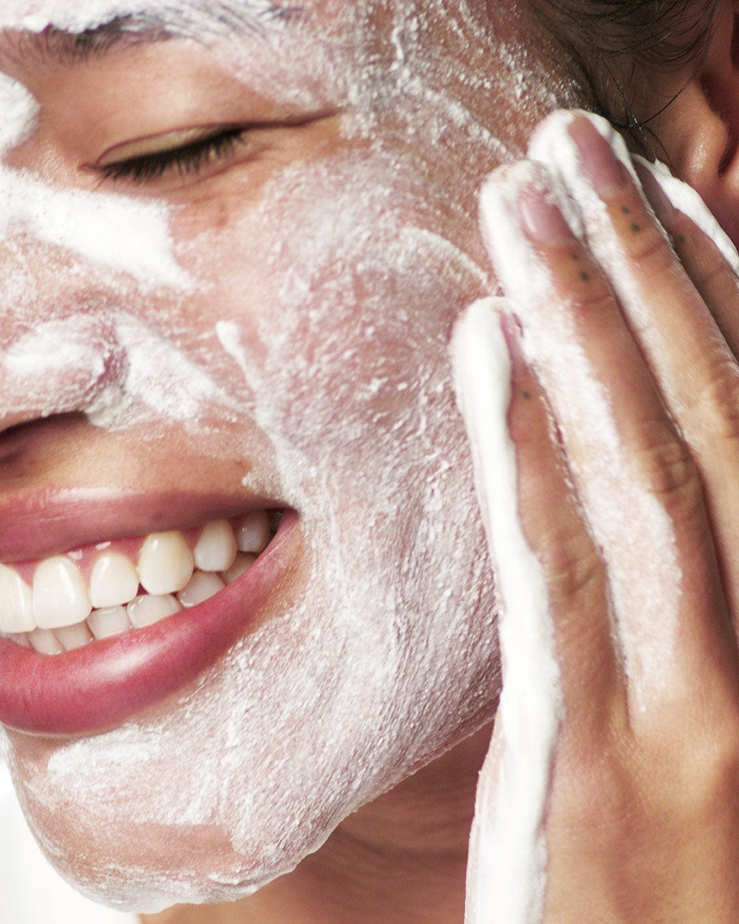 Frau lächelt und streicht mit ihrer Hand über ihr Gesicht, um ein weißes Peeling einzumassieren.