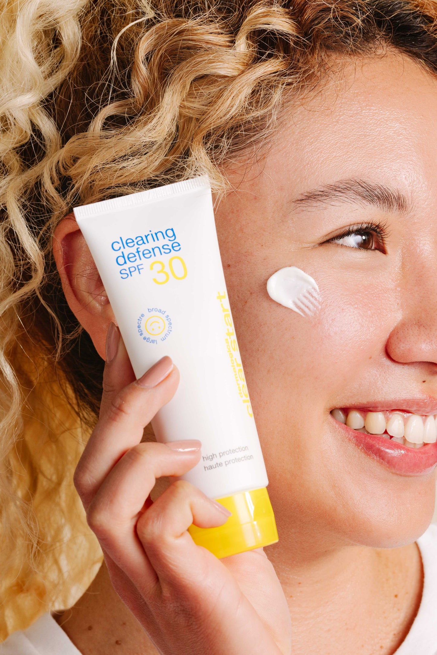 Eine Frau hält eine Tube Clearing Defense SPF30 an ihr Gesicht und tupft etwas davon auf ihre Wange – perfekt für fettige Haut und bietet Breitband-UV-Schutz.