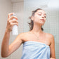 Frau in einem blauen Handtuch sprüht Produkt aus einer weißen Flasche auf ihr Gesicht.