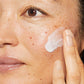 Nahaufnahme des Gesichts einer Person, die Hautpflegecreme auf die Wange aufträgt.