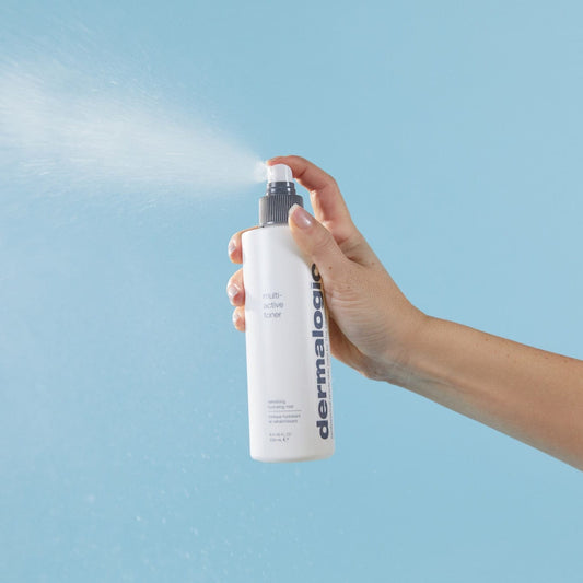 Hand hält Dermalogica Multi-Active Toner Sprayflasche mit sprühendem Nebel vor blauem Hintergrund.
