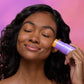 Eine Frau mit geschlossenen Augen trägt den Golden Hour Hydrating SPF 30 Stick auf ihre Wange auf, wodurch ein glasartiger Hautglanz vor einem rosa und violetten Hintergrund zum Vorschein kommt.