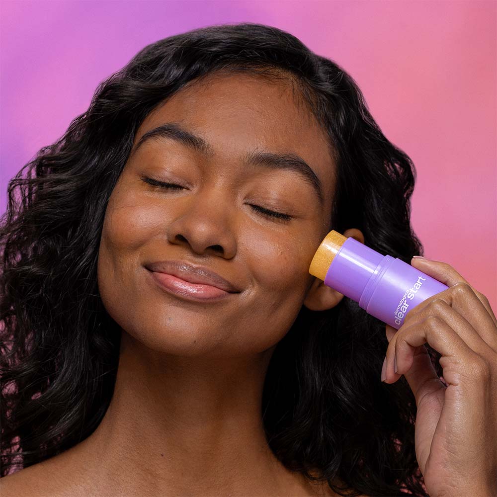 Eine Frau mit geschlossenen Augen trägt den Golden Hour Hydrating SPF 30 Stick auf ihre Wange auf, wodurch ein glasartiger Hautglanz vor einem rosa und violetten Hintergrund zum Vorschein kommt.