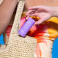 Eine Person steckt einen violetten Hautpflegestift mit der Aufschrift „Golden Hour Hydrating SPF 30 Stick“ in eine gewebte beige Tasche und trägt dabei ein orange-gelbes Oberteil mit Blumenmuster.