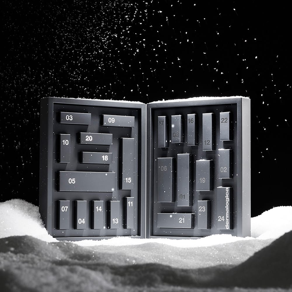 Ein rechteckiger Dermalogica Premium-Adventskalender 2026 mit nummerierten Fächern steht offen und aufrecht im Schnee, während vor schwarzem Hintergrund Schneeflocken fallen – eine ideale Möglichkeit, Ihre Hautpflege-Routine während der festlichen Jahreszeit auf ein neues Niveau zu heben.