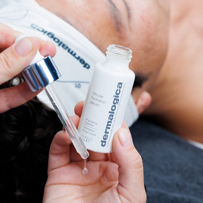 Nahaufnahme von Händen, die eine Pipette und eine Flasche Circular Hydration Serum halten. Im Hintergrund liegt eine Person mit einem Stirnband.