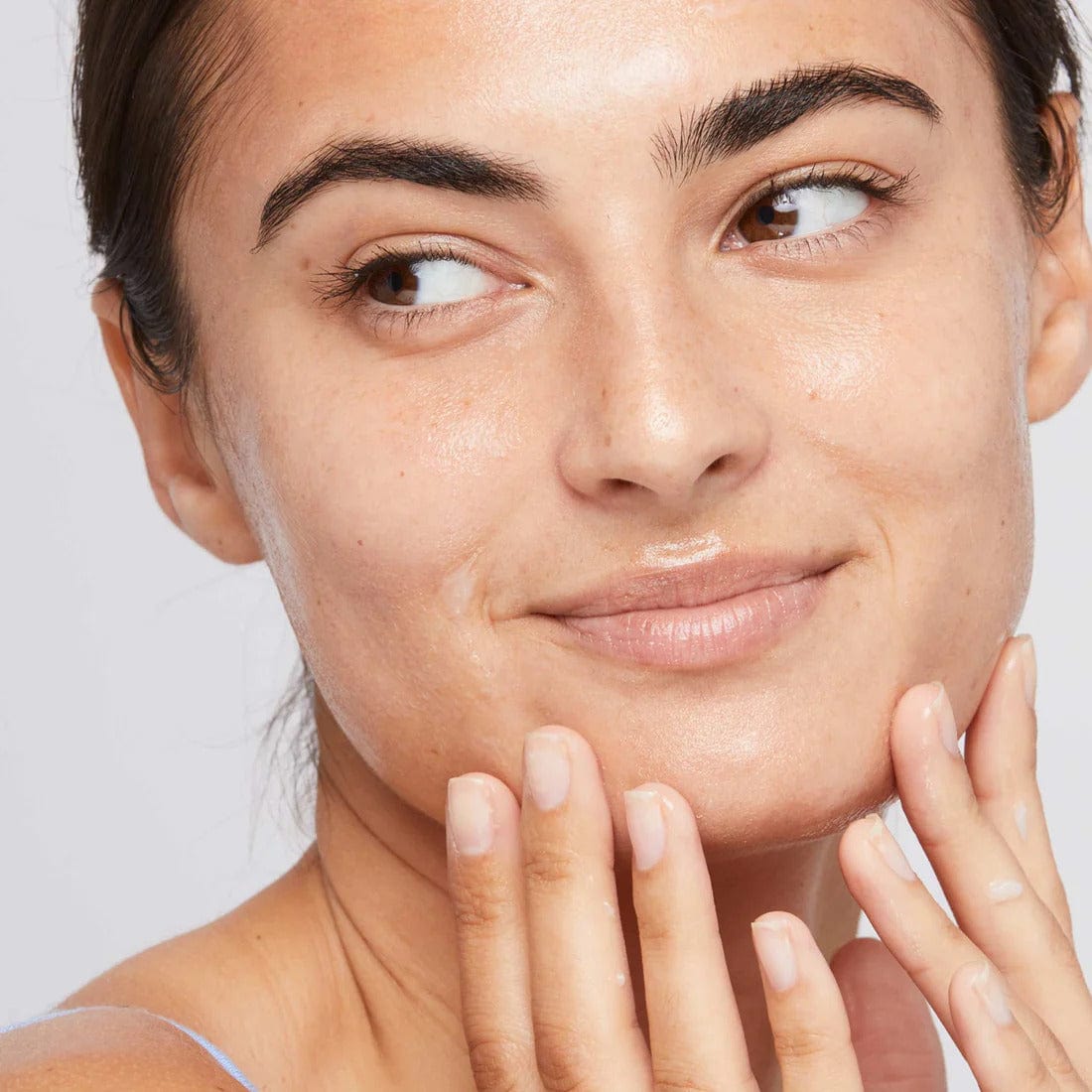 Eine Person mit reiner Haut lächelt, während sie ihr Gesicht berührt, und zeigt eine Nahaufnahme ihrer Hautpflegeroutine mit Calm Water Gel | Feuchtigkeitspflege. Heller Hintergrund, natürlicher Ausdruck.