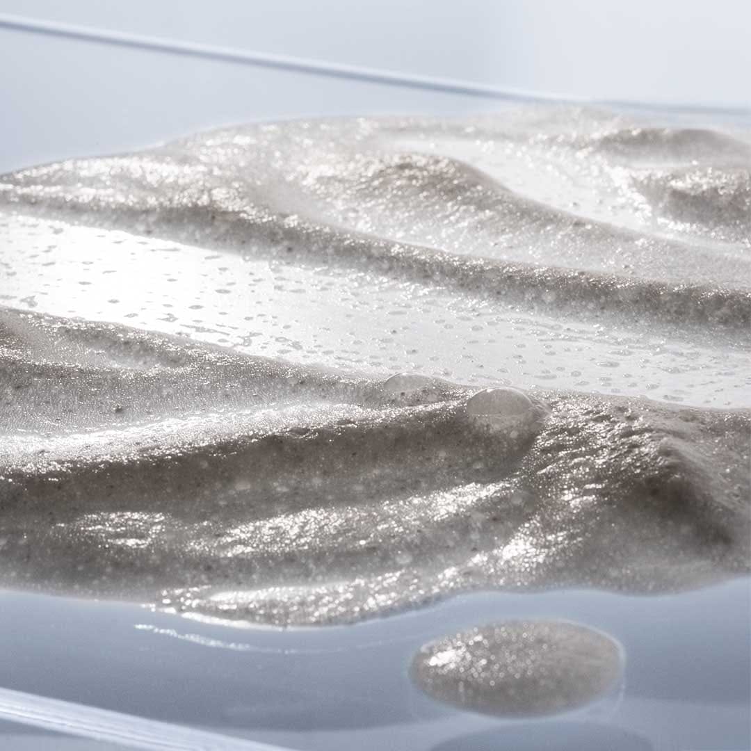 Eine Nahaufnahme des dicken, sprudelnden, gräulichen Daily Microfoliant, der auf einer transparenten Oberfläche aufgetragen wird und perfekt zur Verfeinerung der Hautstruktur geeignet ist.