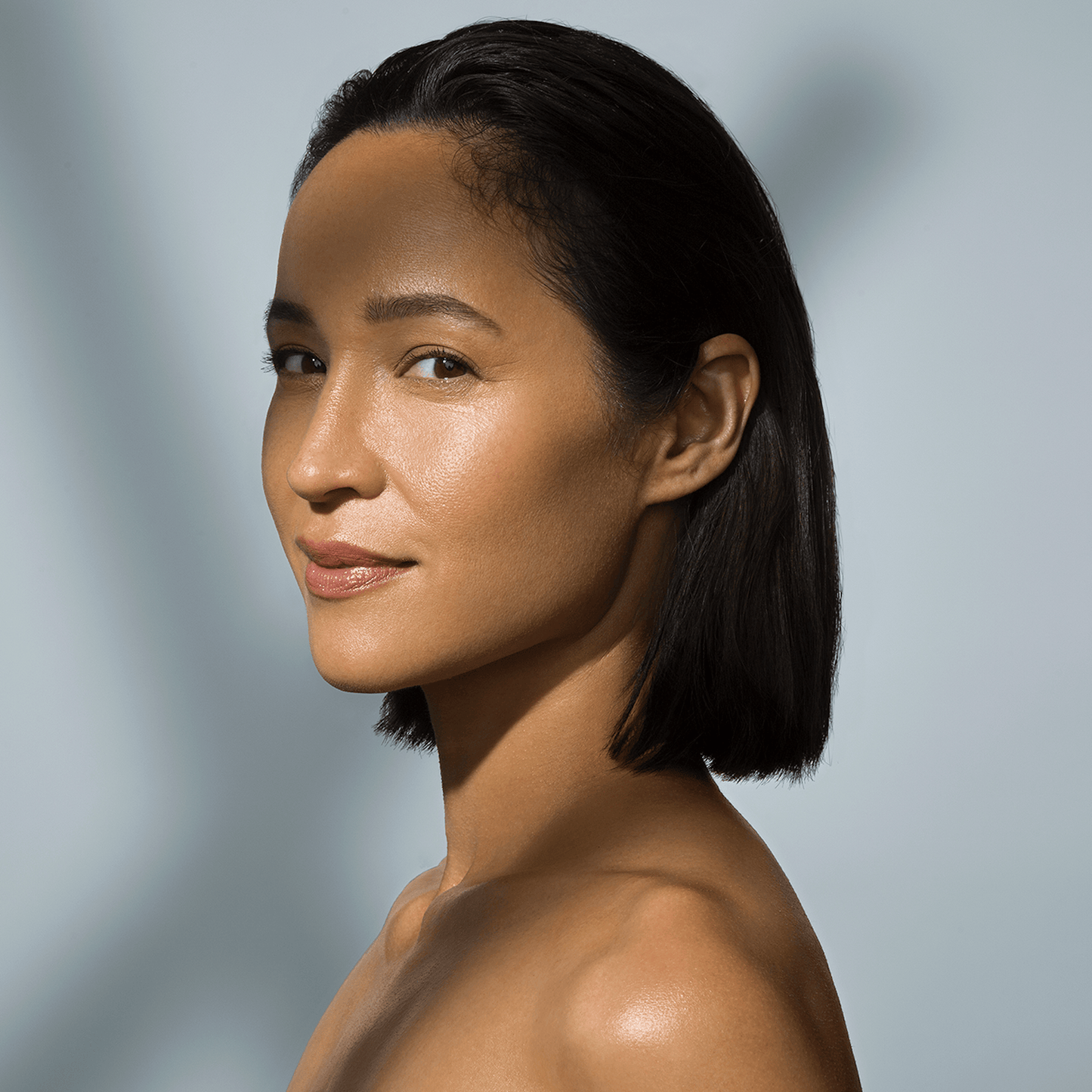 Eine Frau mit kurzem, dunklem Haar und strahlender Haut blickt vor einem sanft beleuchteten Hintergrund über ihre Schulter - ihr strahlender Teint spiegelt die Vorteile des Retinol Clearing Oil wider.