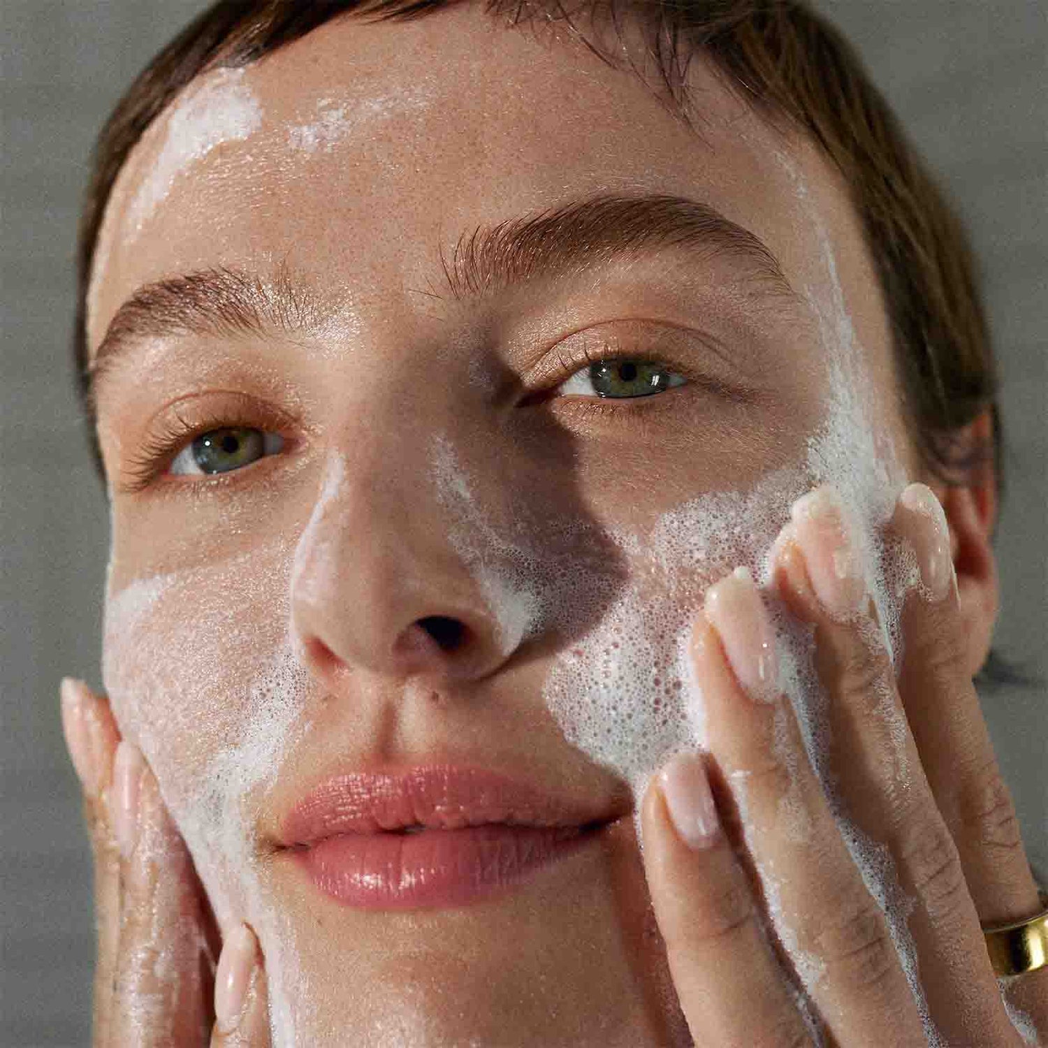 Eine Person mit kurzen Haaren massiert Daily Microfoliant sanft in ihr Gesicht ein und bedeckt Wangen und Kinn mit dem Schaum, um den Teint zu verfeinern (das Hautbild verfeinern).