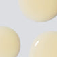 Drei große, glänzende Tropfen einer hellbeigen Flüssigkeit – perfekt für das Biolumin-C Augenserum – sind vor einem hellgrauen Hintergrund zu sehen.