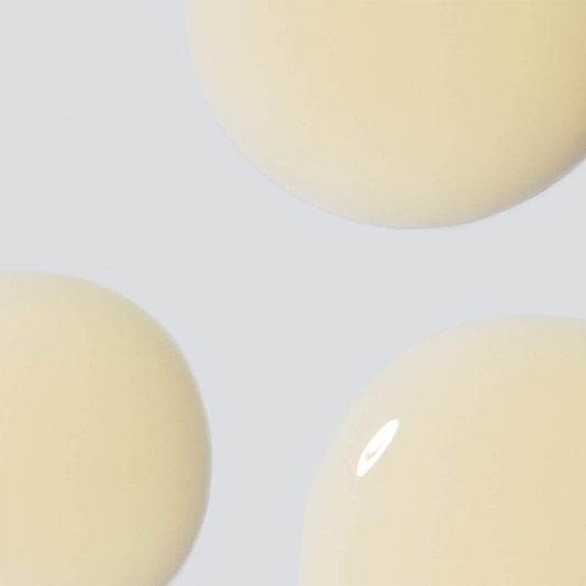 Drei große, glänzende Tropfen einer hellbeigen Flüssigkeit – perfekt für das Biolumin-C Augenserum – sind vor einem hellgrauen Hintergrund zu sehen.