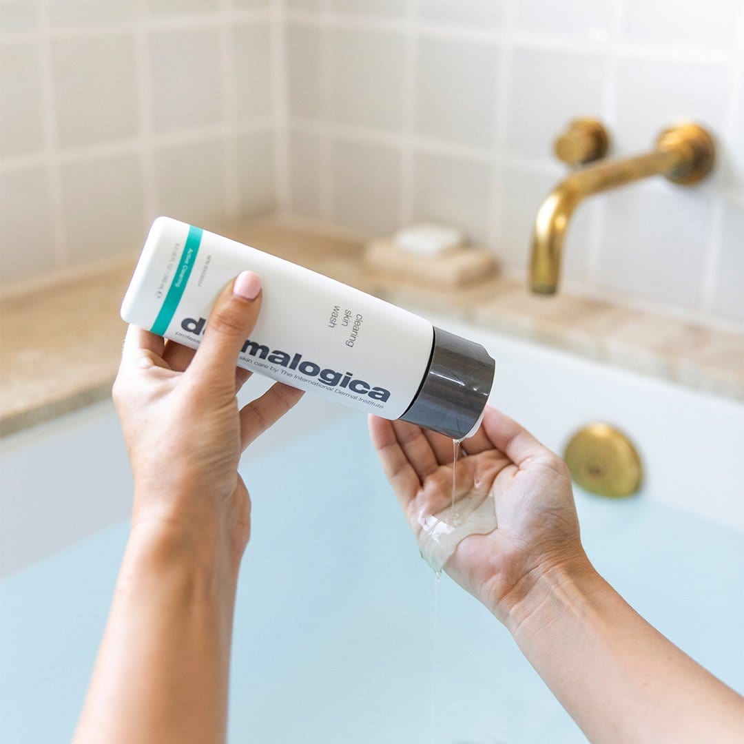Eine Person gießt klares Gel mit reinigendem Schaum aus einer weißen Clearing Skin Wash-Flasche in ihre Hand über einer Badewanne mit Wasser und einem goldenen Wasserhahn.