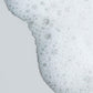 Eine Nahaufnahme von Clearing Skin Wash mit weißen Seifenblasen und Schaum auf einer glatten, hellgrauen Oberfläche.