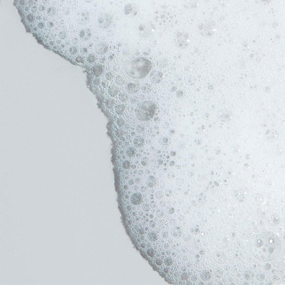 Eine Nahaufnahme von Clearing Skin Wash mit weißen Seifenblasen und Schaum auf einer glatten, hellgrauen Oberfläche.