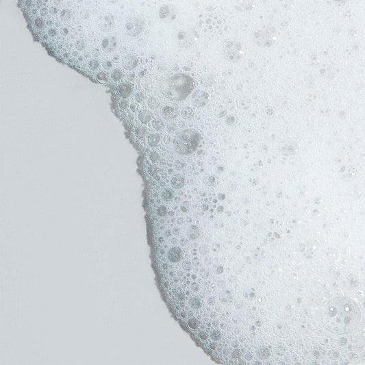 Eine Nahaufnahme von Clearing Skin Wash mit weißen Seifenblasen und Schaum auf einer glatten, hellgrauen Oberfläche.