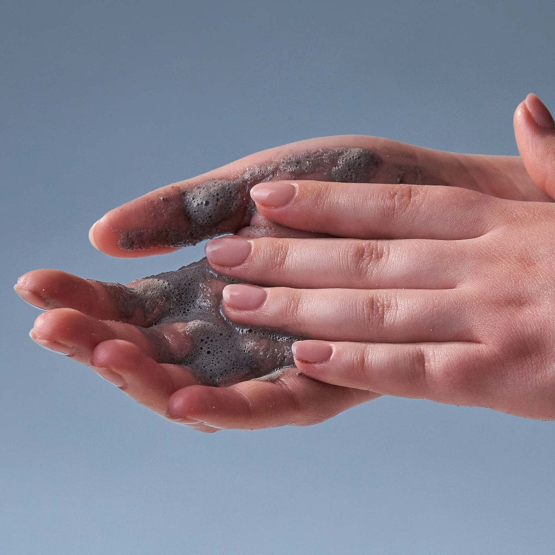 Zwei Hände mit kurzen, blassrosa Nägeln reiben sich mit Daily Superfoliant-Schaum – der die Kollagenproduktion unterstützt – vor einem schlichten blauen Hintergrund aneinander.