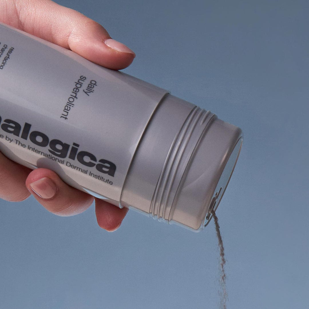 Eine Hand gießt Daily Superfoliant aus einer grauen Flasche vor einem schlichten blauen Hintergrund, um Hautschäden zu bekämpfen und für glattere Haut zu sorgen.