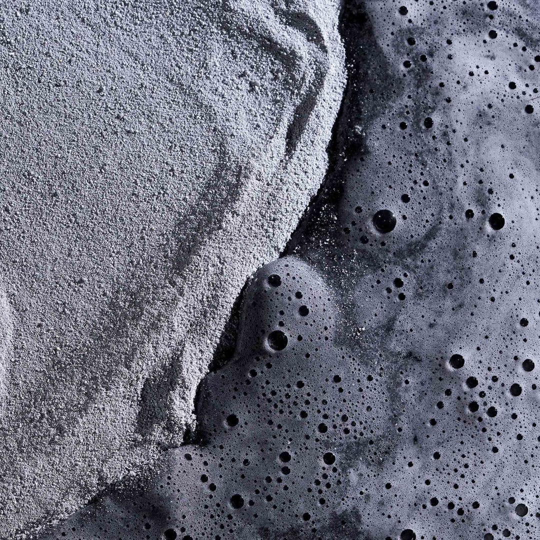 Nahaufnahme des feinen grauen Daily Superfoliant-Puders, der mit einer dunklen, sprudelnden Flüssigkeit verschmilzt, was auf eine erhöhte Kollagenproduktion ohne Risiko von Hautschäden hindeutet.