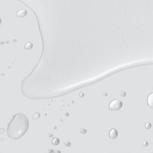 Klare Wassertropfen und eine kleine Pfütze verteilen sich auf einer glatten, hellgrauen Oberfläche und erinnern an die frische Feuchtigkeitsversorgung durch Retinol Clearing Oil.