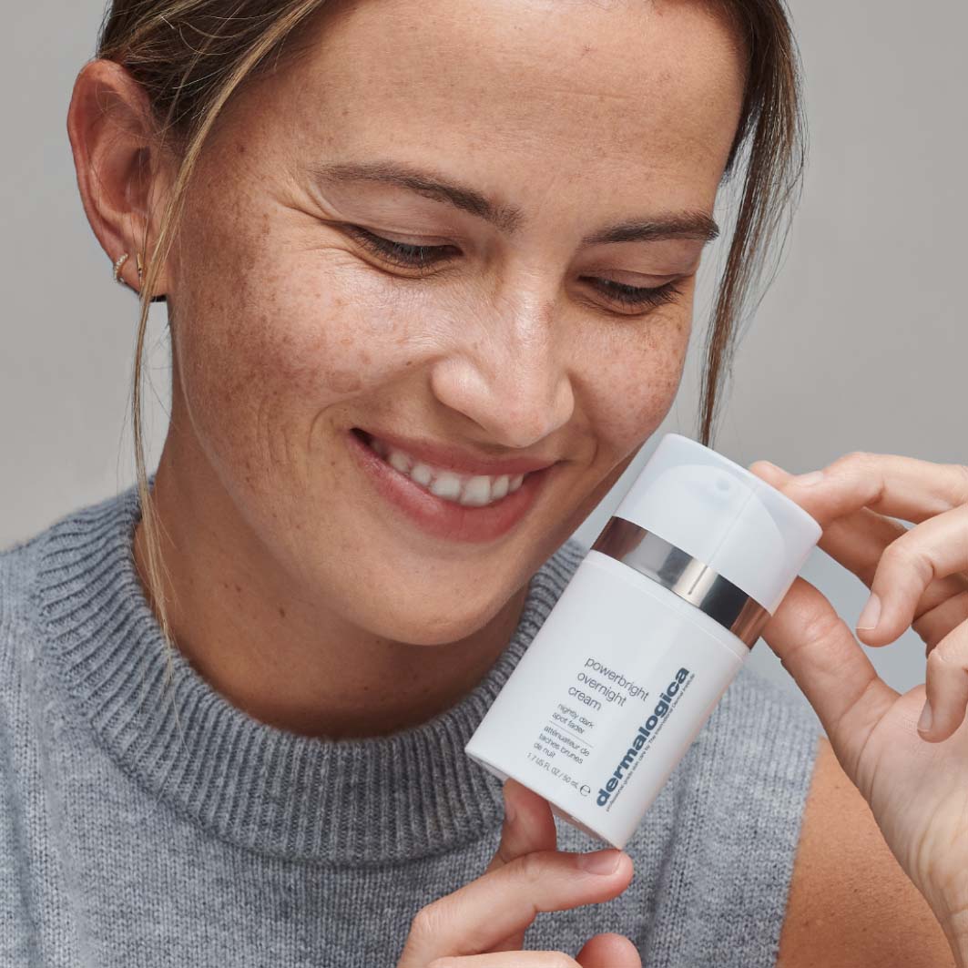 Lächelnde Frau hält ein Produkt von Dermalogica, die "PowerBright Overnight Cream", nah an ihr Gesicht.