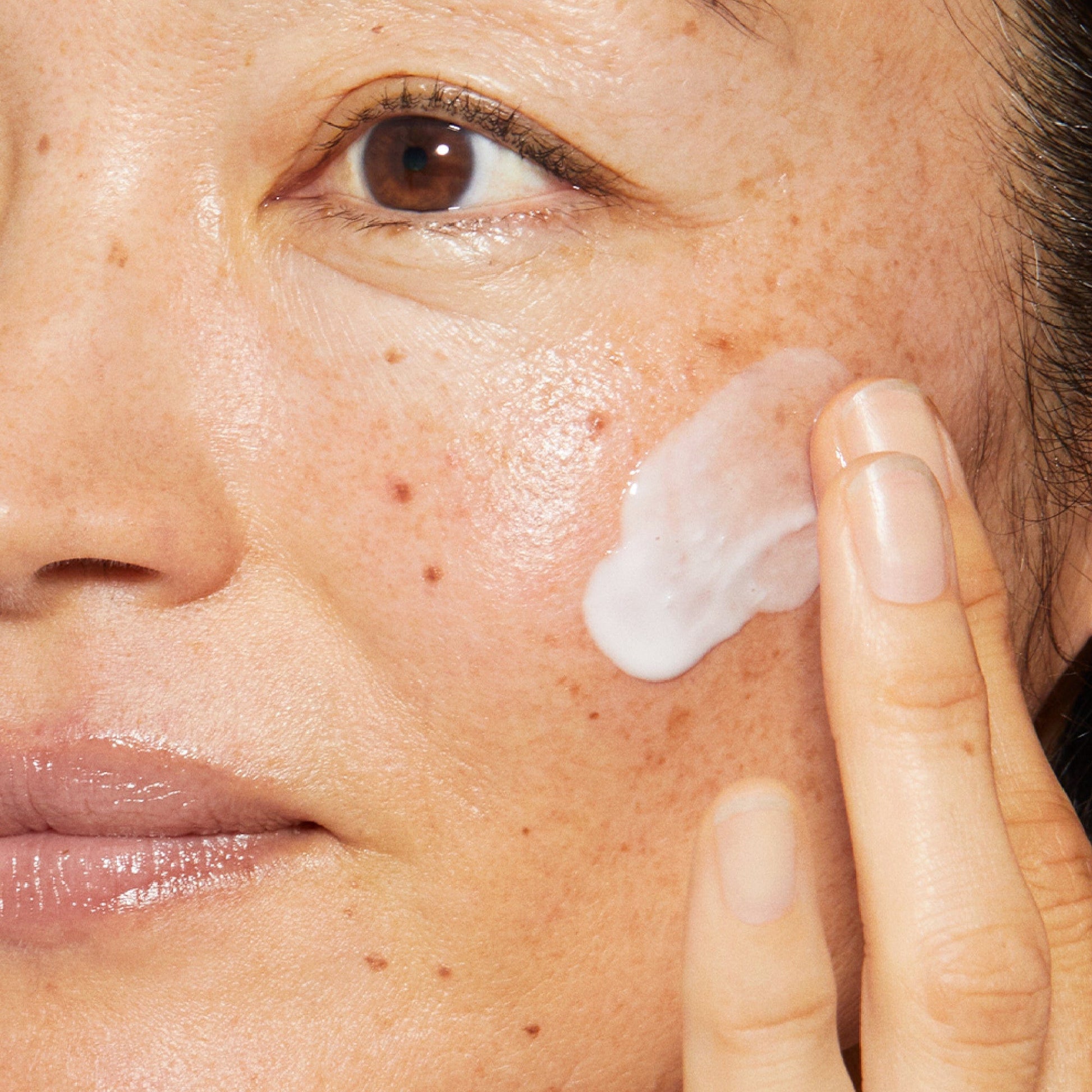 Nahaufnahme des Gesichts einer Person, die Hautpflegecreme auf die Wange aufträgt.