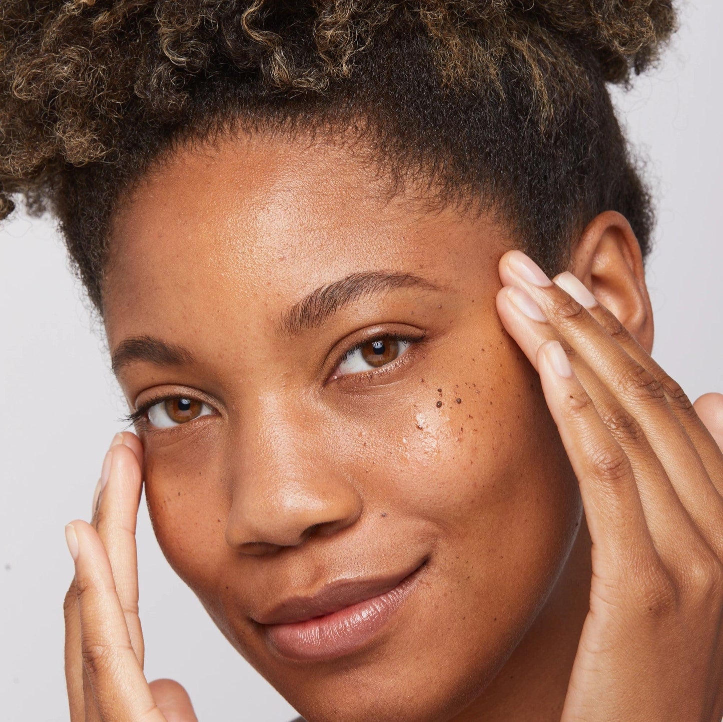 Eine Frau mit Afrohaar berührt sanft ihr Gesicht mit ihren Händen und profitiert dabei von den pigmentausgleichenden Eigenschaften des Biolumin-C Augenserums 15 ml | Augenpflege.