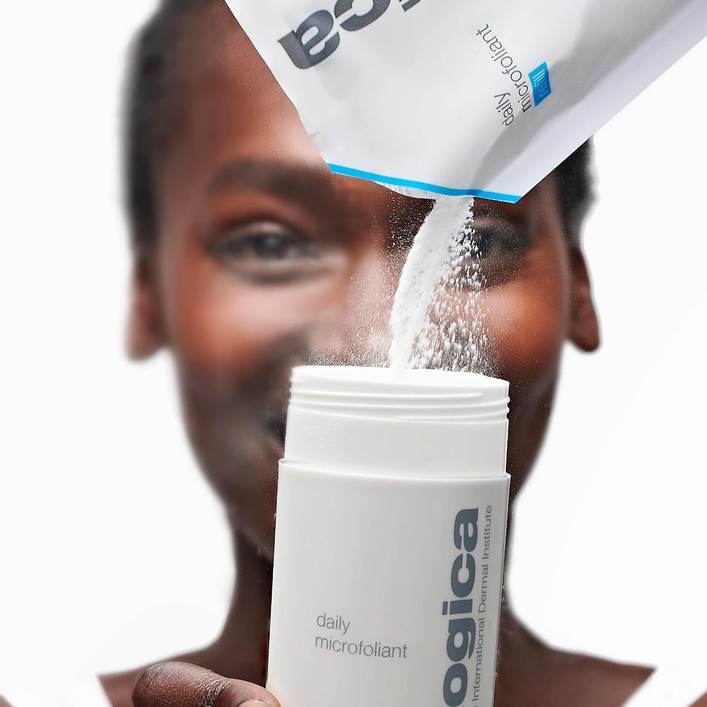 Eine Person hält eine Flasche "Dermalogica Daily Microfoliant" in der Hand, aus der ein Pulver auf einen weiteren Teil des Gesichts der Person zu fallen scheint.