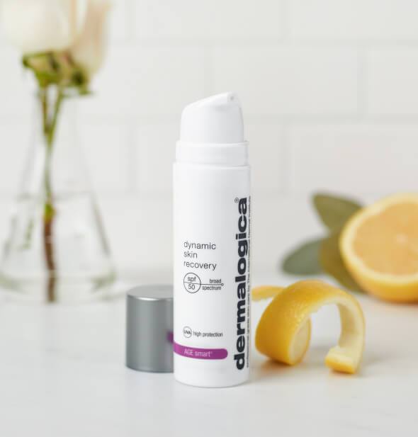 Flasche Dermalogica Dynamic Skin Recovery SPF50 mit offenem Deckel neben Zitronenscheiben und einer Rose im Hintergrund.