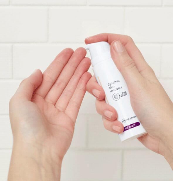 Hände halten eine Flasche Dermalogica Dynamic Skin Recovery SPF50 Hautcreme vor einem weißen Hintergrund.